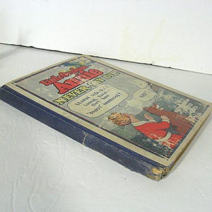 Pu&ograve; includere: Un libro vintage blu intitolato "Little Orphan Annie Never Can Die" con un'illustrazione a fumetti di una donna e un cane sulla copertina.