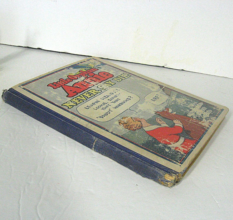 Pu&ograve; includere: Un libro vintage blu intitolato "Little Orphan Annie Never Can Die" con un'illustrazione a fumetti di una donna e un cane sulla copertina.