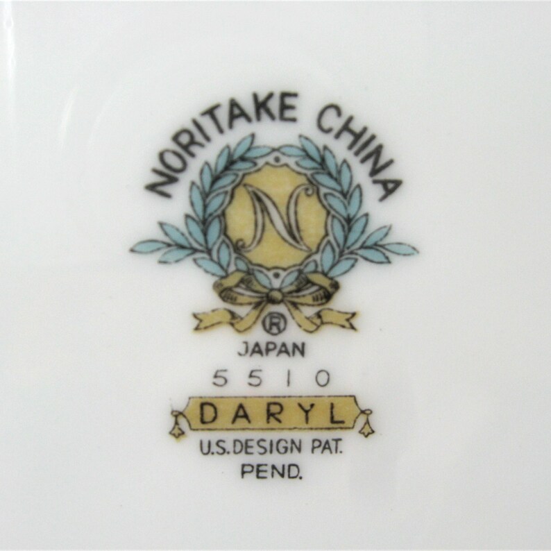 Puede incluir: Un plato de cer&aacute;mica blanco con un logotipo de Noritake China azul y dorado. El logotipo presenta una corona con la letra N en el centro. El texto "Japan 5510 Daryl U.S. Design Pat. Pend." est&aacute; impreso debajo del logotipo.