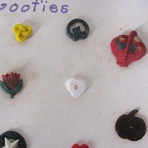 May include: A collection of assorted decorative buttons including a yellow knot, a black ring, a red basket, a red and green tulip, a white heart, a dark brown apple, and a black and white star, arranged on a surface with the text Goofies.