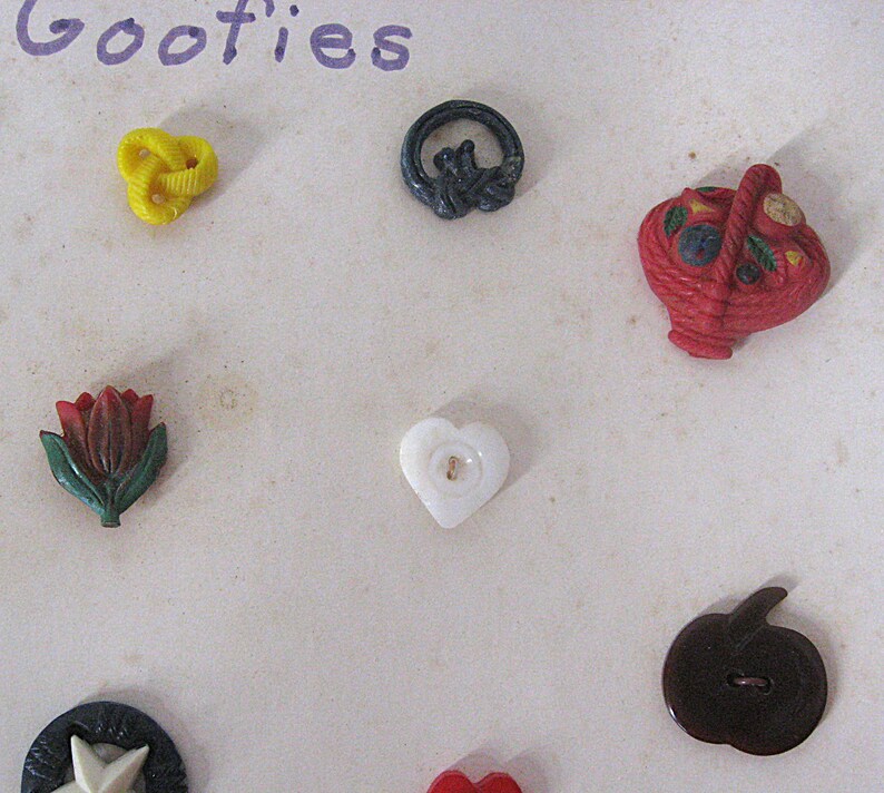 May include: A collection of assorted decorative buttons including a yellow knot, a black ring, a red basket, a red and green tulip, a white heart, a dark brown apple, and a black and white star, arranged on a surface with the text Goofies.