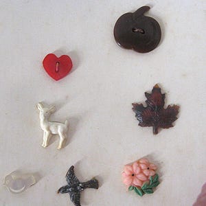 May include: A collection of seven assorted buttons featuring a dark brown apple, a red heart, a white deer, a dark brown maple leaf, a translucent bird, a dark grey bird in flight, and a cluster of pink flowers with green leaves.