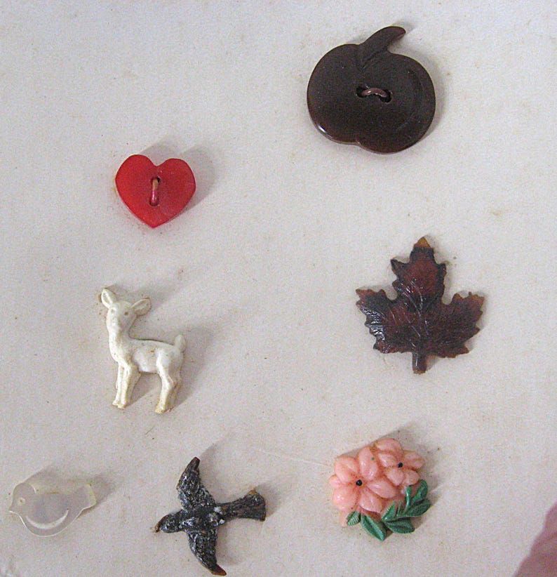 May include: A collection of seven assorted buttons featuring a dark brown apple, a red heart, a white deer, a dark brown maple leaf, a translucent bird, a dark grey bird in flight, and a cluster of pink flowers with green leaves.