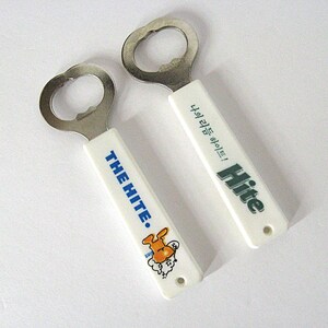May include: Two bottle openers with white handles and silver metal opener heads. One handle has the text "THE HITE." in blue, with a cartoon character. The other has the text "Hite" in green. The background is white.