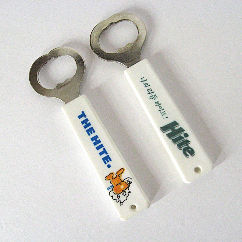 May include: Two bottle openers with white handles and silver metal opener heads. One handle has the text "THE HITE." in blue, with a cartoon character. The other has the text "Hite" in green. The background is white.