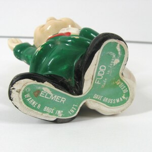 May include: A green ceramic figurine of Elmer Fudd, a character from the Looney Tunes cartoon series. The figurine is wearing a green jacket and a red bow tie. The figurine is sitting down with its arms raised. The figurine is made by Dave Grossman and is dated 1971.