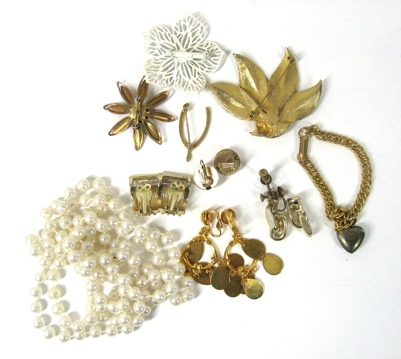 K&ouml;nnte beinhalten: Eine Sammlung von Vintage-Schmuck, darunter eine Perlenkette, goldfarbene Ohrringe, ein goldfarbenes Armband und mehrere Broschen. Die Broschen haben die Form einer Blume, eines Blattes und eines Sterns.