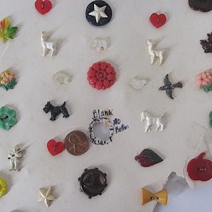 May include: A collection of assorted decorative buttons featuring various shapes including red hearts, maple leaves, animals like deer and dogs, flowers, stars, and a sailboat. A copper penny is included for scale next to a handwritten note.