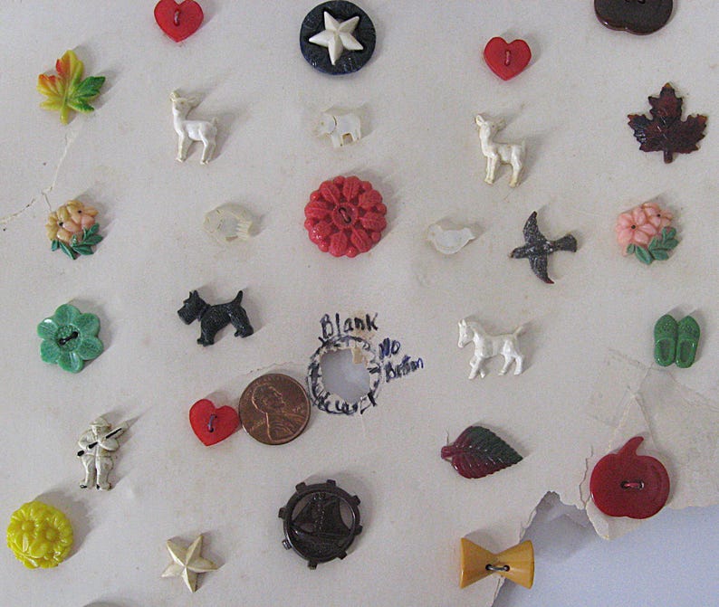 May include: A collection of assorted decorative buttons featuring various shapes including red hearts, maple leaves, animals like deer and dogs, flowers, stars, and a sailboat. A copper penny is included for scale next to a handwritten note.