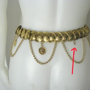 May include: Gold-toned belt featuring a series of circular medallions with embossed designs. The belt has dangling chains and smaller coin-shaped charms. The belt is displayed on a mannequin.