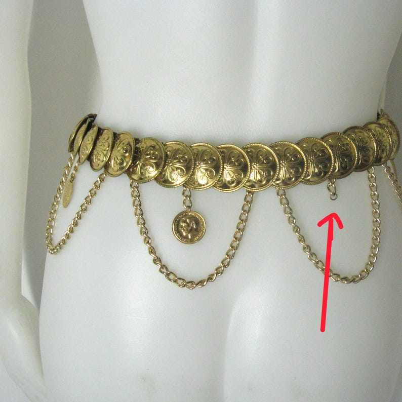 May include: Gold-toned belt featuring a series of circular medallions with embossed designs. The belt has dangling chains and smaller coin-shaped charms. The belt is displayed on a mannequin.