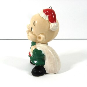 May include: A ceramic Christmas ornament of a bald-headed character wearing a green suit, a red Santa hat, and carrying a white sack.