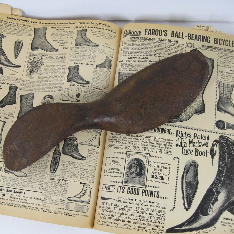May include: A rusty metal shoe stretcher with a curved handle. The stretcher is resting on an open book with a vintage advertisement for shoes. The ad features the text "Richy's Patent Julia Marlowe Lace Boot" and "A Few of Its Good Points."