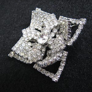 May include: A silver-tone brooch featuring a large, clear rhinestone centre surrounded by smaller rhinestones. The brooch is shaped like a bow.