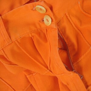 May include: Close-up of orange fabric with a zipper and two buttons. The fabric is pleated and has a textured surface.