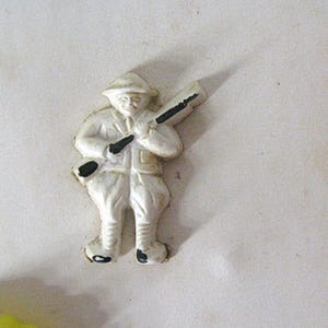 May include: A small, white, molded plastic figurine of a soldier wearing a helmet and uniform, carrying a rifle over the shoulder. The figure features black painted accents on the rifle barrel, stock, and boots, with a flat back design.