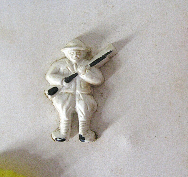 May include: A small, white, molded plastic figurine of a soldier wearing a helmet and uniform, carrying a rifle over the shoulder. The figure features black painted accents on the rifle barrel, stock, and boots, with a flat back design.