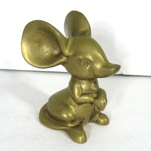 May include: A solid brass figurine of a mouse with large ears. The mouse is sitting with its paws folded in front of it. The figurine is a golden color.