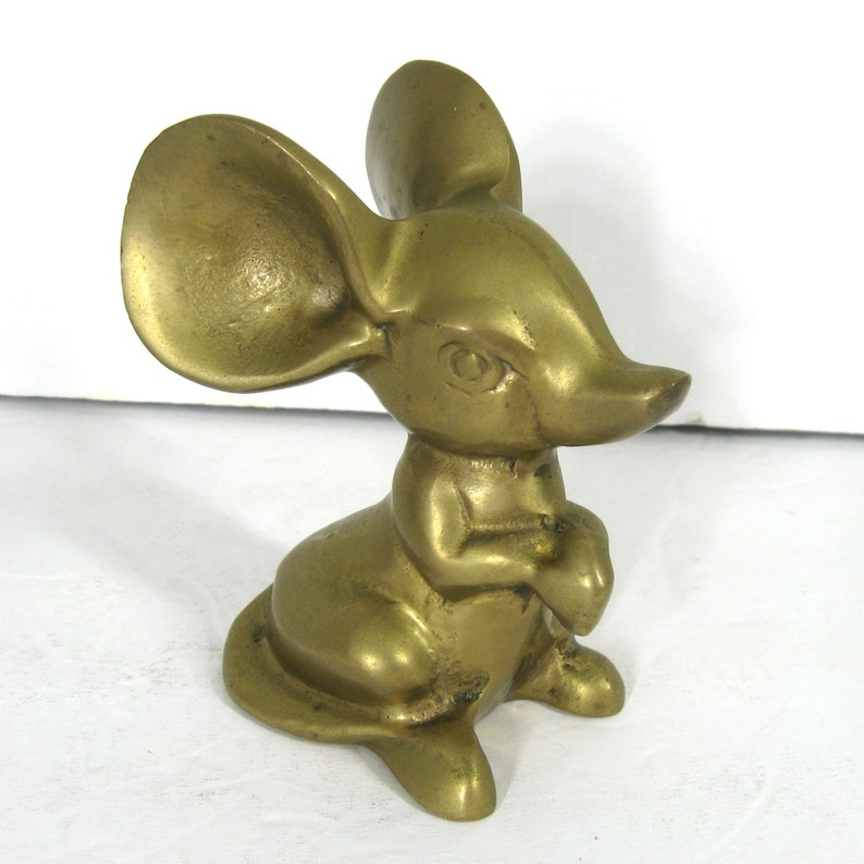 May include: A solid brass figurine of a mouse with large ears. The mouse is sitting with its paws folded in front of it. The figurine is a golden color.