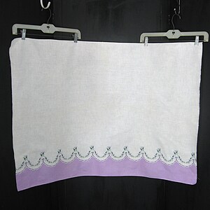 May include: A white linen towel with a lavender scalloped border. The border is decorated with small embroidered floral accents in shades of green and purple. The towel is hanging from a plastic hanger.