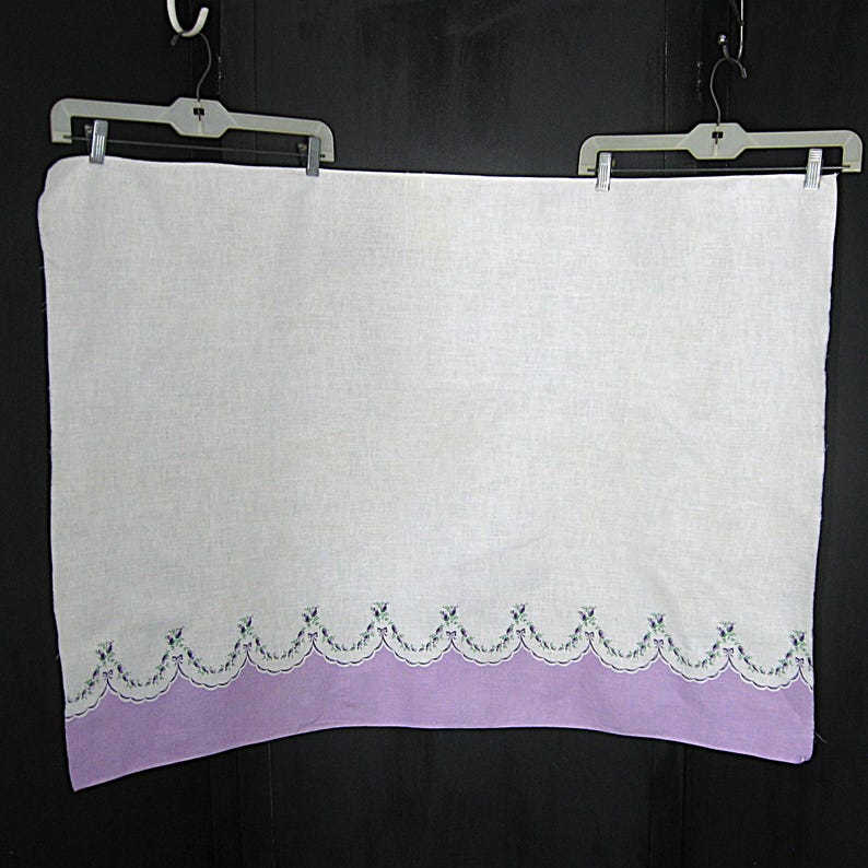 May include: A white linen towel with a lavender scalloped border. The border is decorated with small embroidered floral accents in shades of green and purple. The towel is hanging from a plastic hanger.