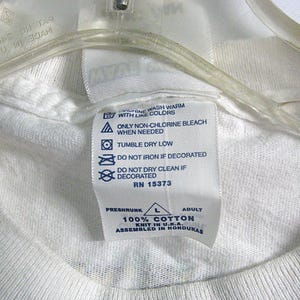 May include: White cotton t-shirt tag with blue text and symbols. The tag includes washing instructions, size, and fabric content. The text reads "Machine wash warm with like colors. Only non-chlorine bleach when needed. Tumble dry low. Do not iron if decorated. Do not dry clean if decorated. RN 15373. Preshrunk 100% Cotton Knit in U.S.A. Assembled in Honduras."