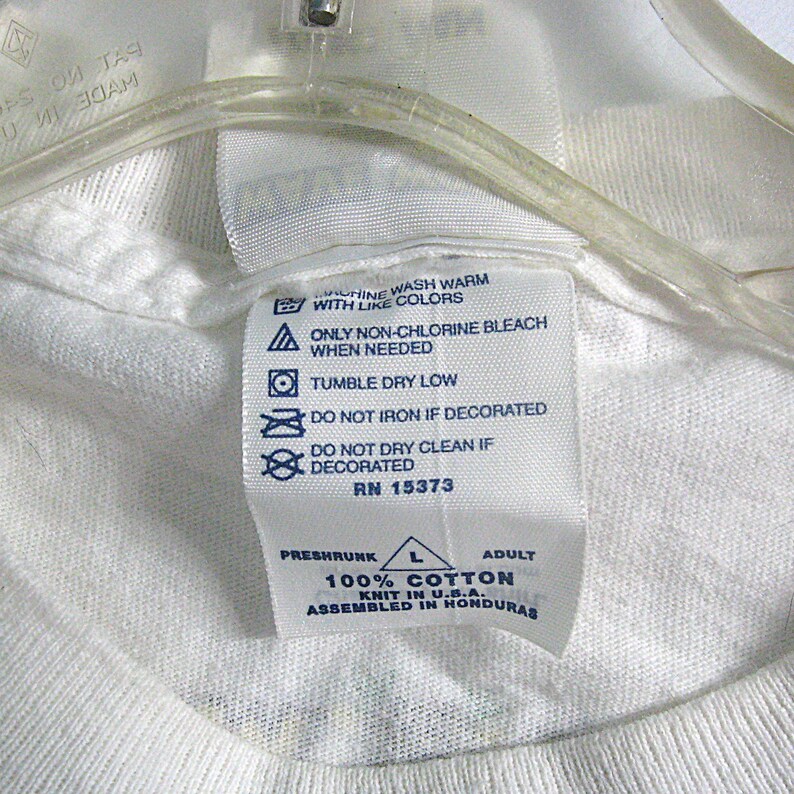 May include: White cotton t-shirt tag with blue text and symbols. The tag includes washing instructions, size, and fabric content. The text reads "Machine wash warm with like colors. Only non-chlorine bleach when needed. Tumble dry low. Do not iron if decorated. Do not dry clean if decorated. RN 15373. Preshrunk 100% Cotton Knit in U.S.A. Assembled in Honduras."