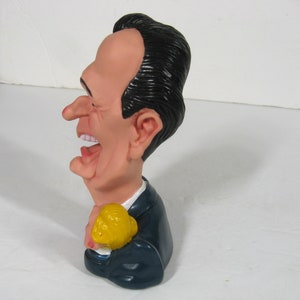 May include: A caricature figurine of a man with black hair, a blue suit, and a yellow object in his hand. The figurine is made of plastic and has a humorous expression on its face.