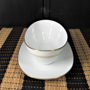 K&ouml;nnte beinhalten: Wei&szlig;e Porzellan-So&szlig;enk&auml;nnchen mit goldener Verzierung und geometrischem Muster. Die Kanne steht auf einer wei&szlig;en Untertasse mit goldener Verzierung.