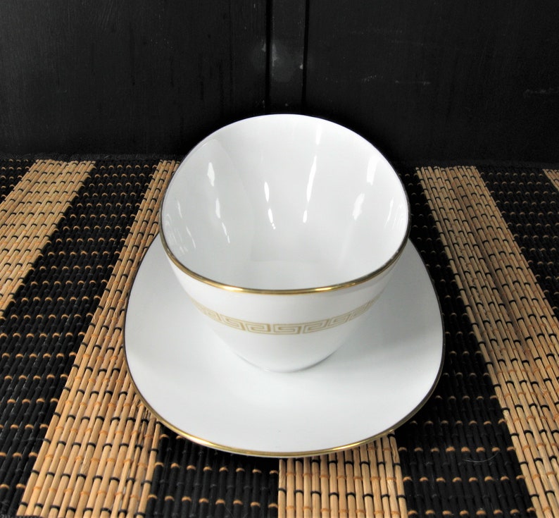K&ouml;nnte beinhalten: Wei&szlig;e Porzellan-So&szlig;enk&auml;nnchen mit goldener Verzierung und geometrischem Muster. Die Kanne steht auf einer wei&szlig;en Untertasse mit goldener Verzierung.