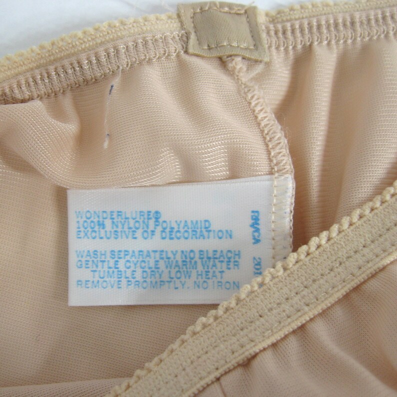 May include: A white tag with blue text attached to a beige garment. The tag reads "Wonderlure 100% Nylon Polyamid Exclusive of Decoration Wash Separately No Bleach Gentle Cycle Warm Water Tumble Dry Low Heat Remove Promptly. No Iron" and "Praca 201".