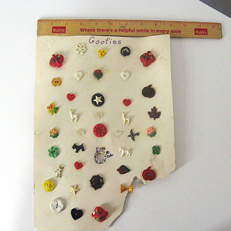 May include: A collection of assorted small decorative buttons labeled Goofies on a rectangular card, displayed with a wooden ruler measuring 12 inches at the top. The buttons feature various shapes including hearts, animals, leaves, and flowers in colors like red, white, yellow, green, and black.