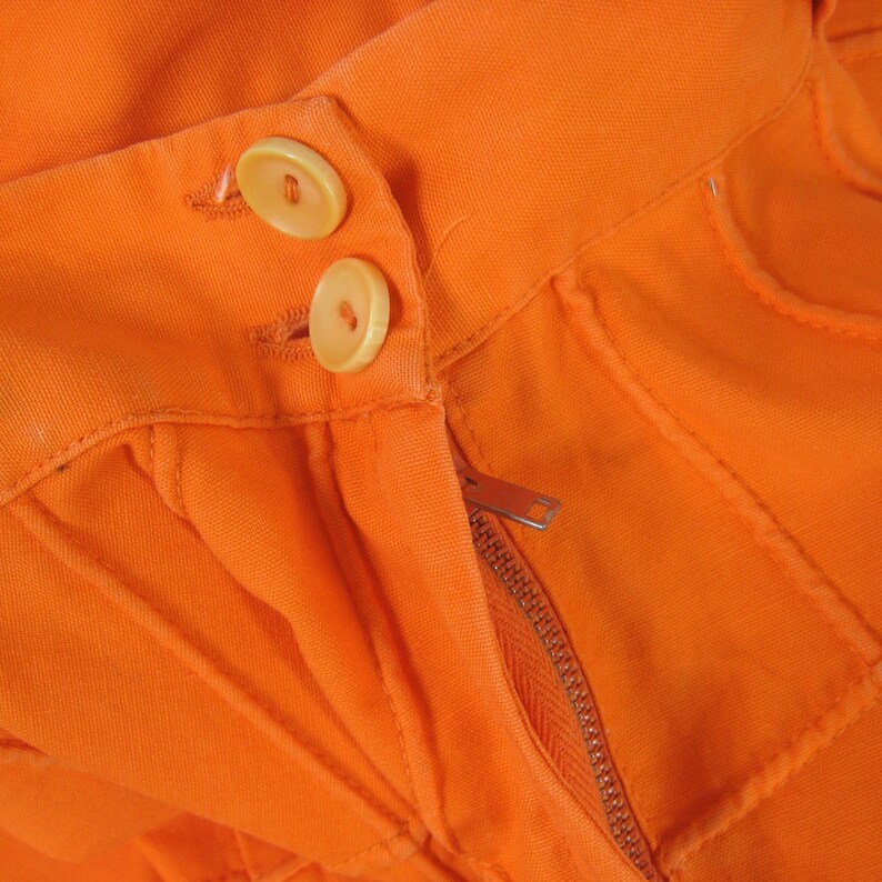 May include: Close-up of the waistband of a pair of orange pants with two orange buttons and a zipper.