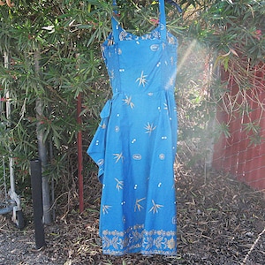 May include: A blue sundress with a floral and bamboo leaf pattern. The dress has a fitted bodice, adjustable straps, and a flowing skirt. Gold accents are visible on the hem and bodice. The dress is hanging outside on a hanger.
