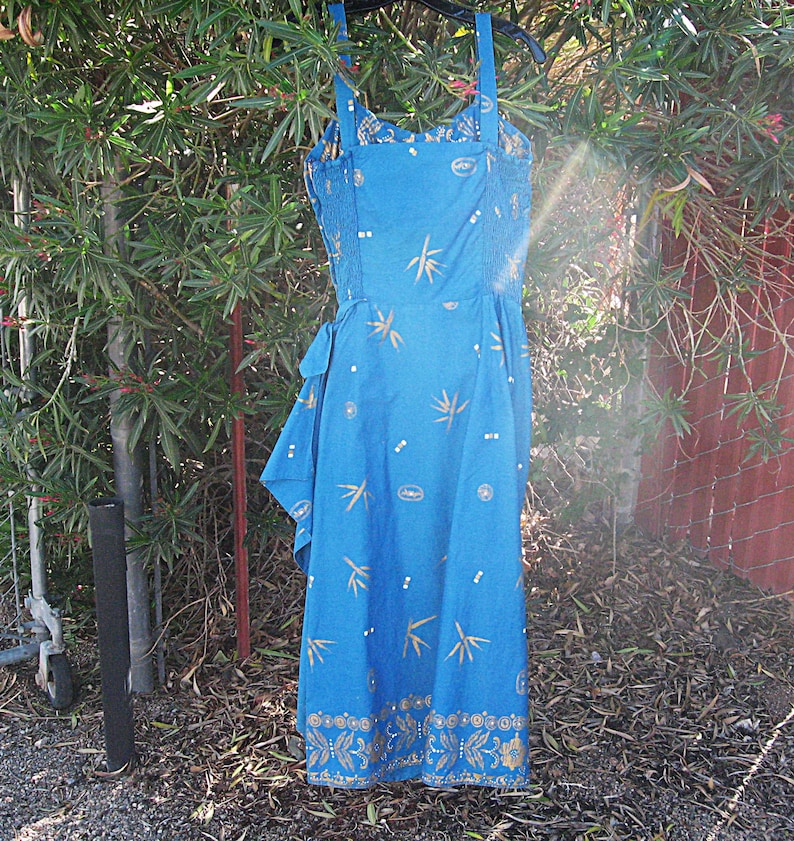 May include: A blue sundress with a floral and bamboo leaf pattern. The dress has a fitted bodice, adjustable straps, and a flowing skirt. Gold accents are visible on the hem and bodice. The dress is hanging outside on a hanger.