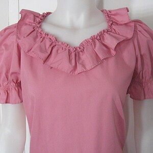 Puede incluir: Una blusa rosa s&oacute;lida con mangas cortas abullonadas con pu&ntilde;os el&aacute;sticos con volantes y un escote ancho y redondeado adornado con un prominente volante de tela fruncida que se extiende alrededor del cuello, creando una apariencia texturizada.