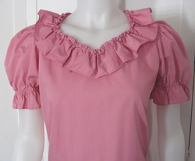 Puede incluir: Una blusa rosa s&oacute;lida con mangas cortas abullonadas con pu&ntilde;os el&aacute;sticos con volantes y un escote ancho y redondeado adornado con un prominente volante de tela fruncida que se extiende alrededor del cuello, creando una apariencia texturizada.