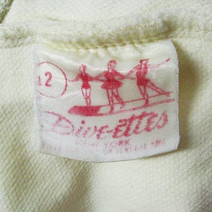 Op de afbeelding: Een wit label met een rode cirkel met het nummer 12. Het label heeft ook een rode afbeelding van drie ballerina's die elkaar vasthouden en de tekst "Dixie-Ettes" in het rood.