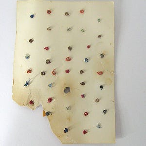 May include: A rectangular, aged, off-white paper card featuring numerous small, colorful metal wire twists or fasteners attached in a grid-like pattern. The paper shows signs of wear, including frayed edges, discoloration, and a small central hole.