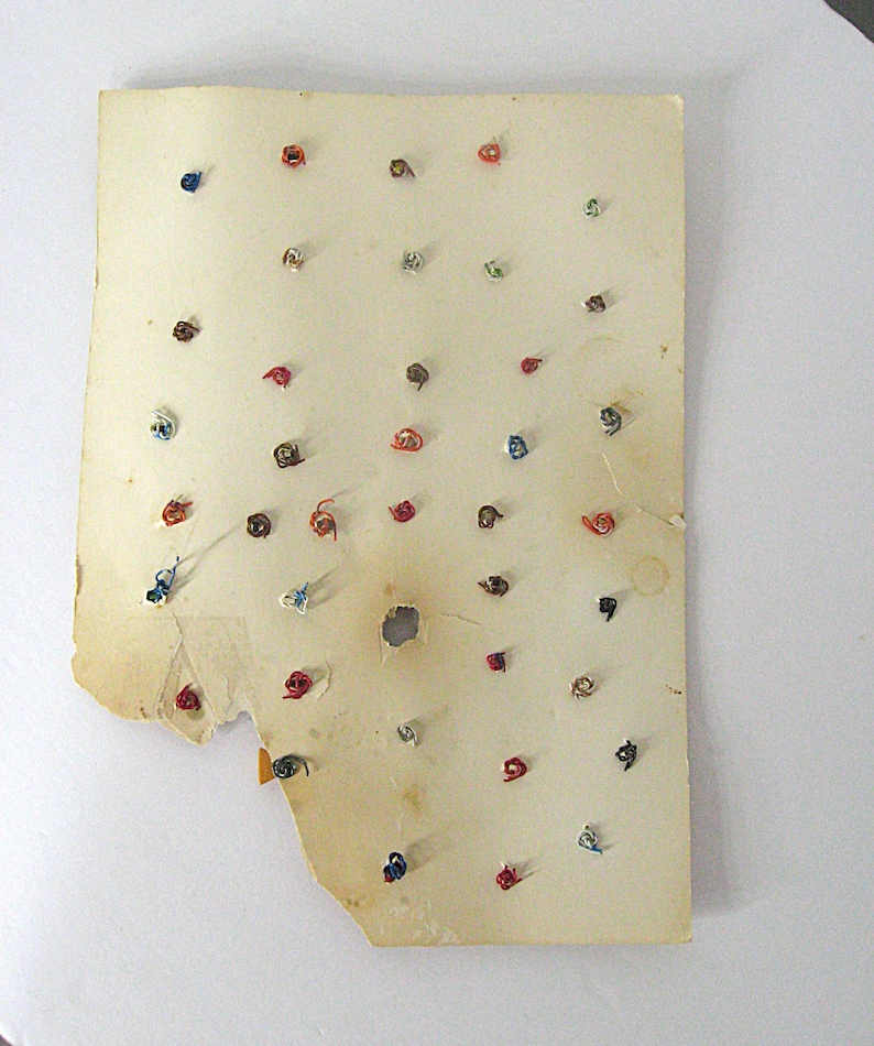 May include: A rectangular, aged, off-white paper card featuring numerous small, colorful metal wire twists or fasteners attached in a grid-like pattern. The paper shows signs of wear, including frayed edges, discoloration, and a small central hole.