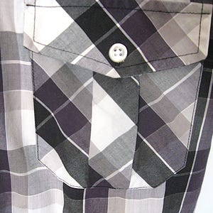 May include: Close-up of a button-down shirt with a plaid pattern in shades of gray, black, and white. The shirt features a pocket on the chest with a white button closure. The fabric has a subtle sheen.