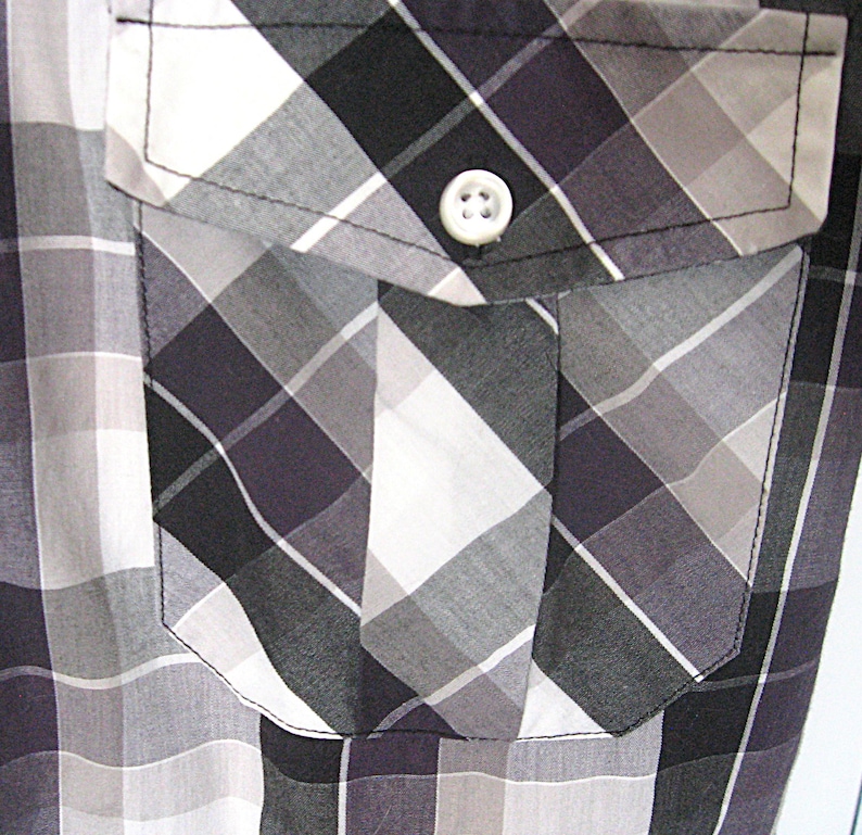 May include: Close-up of a button-down shirt with a plaid pattern in shades of gray, black, and white. The shirt features a pocket on the chest with a white button closure. The fabric has a subtle sheen.