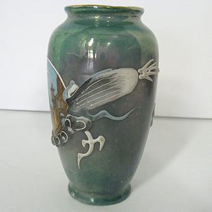 Pode incluir: Um vaso de cer&acirc;mica verde com um desenho de drag&atilde;o. O vaso tem um gradiente verde escuro e cinza com uma borda dourada. O drag&atilde;o &eacute; branco e cinza com detalhes em azul, e um edif&iacute;cio &eacute; vis&iacute;vel na lateral.