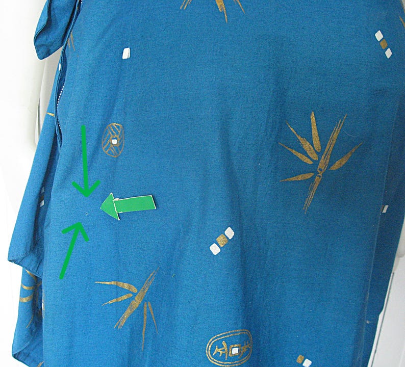 May include: Teal fabric featuring a pattern of gold bamboo-style designs and white and gold geometric shapes. The fabric is part of a garment, with a visible seam and a portion of a sleeve.