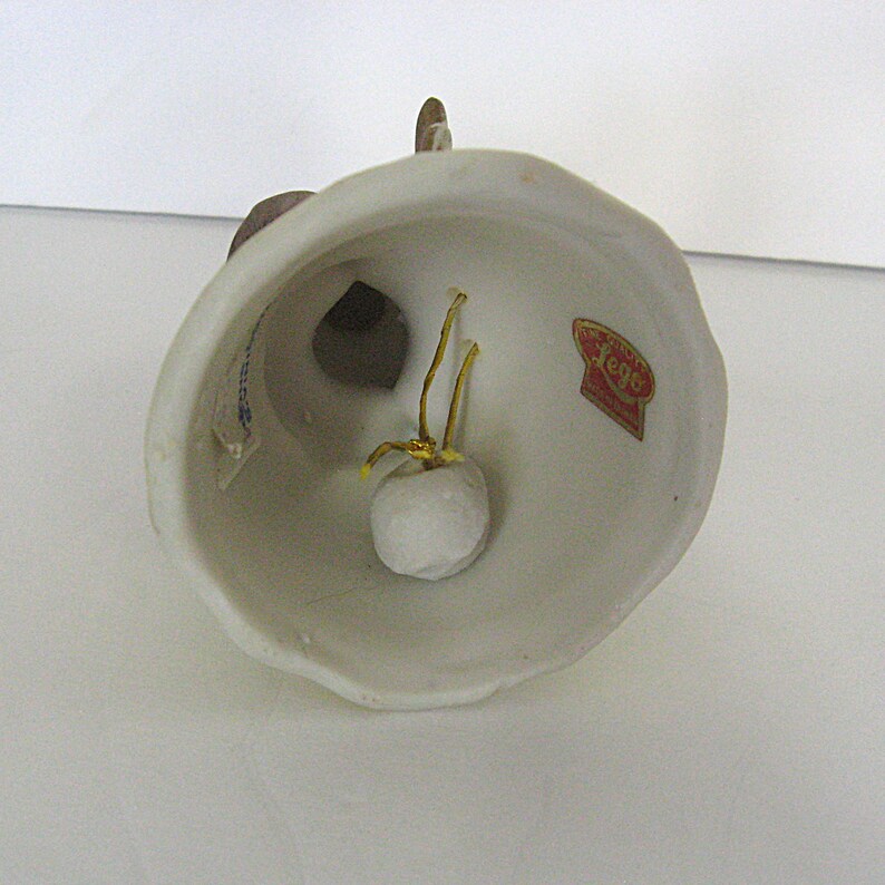 May include: A white ceramic bell with a brown figure on top. The bell has a gold clapper and a small white ball inside. A red and gold sticker is visible on the side.