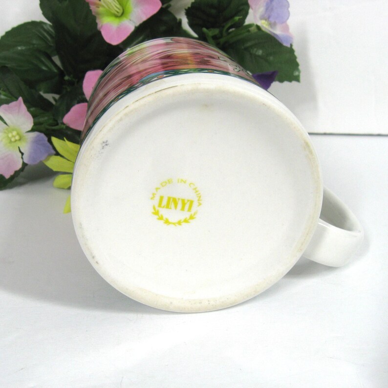 May include: White ceramic mug with a floral design and a yellow "LINYI" logo on the bottom. The logo is surrounded by a wreath and the words "MADE IN CHINA".