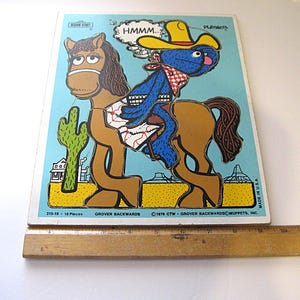 Pode incluir: Um quebra-cabe&ccedil;a de 10 pe&ccedil;as da Sesame Street intitulado Grover Backwards, com um personagem azul de chap&eacute;u de cowboy amarelo montado em um cavalo marrom em um deserto com um cacto. Uma r&eacute;gua de madeira mostra 25,4 cm de largura.