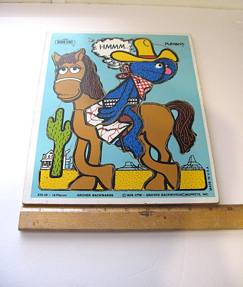 Pode incluir: Um quebra-cabe&ccedil;a de 10 pe&ccedil;as da Sesame Street intitulado Grover Backwards, com um personagem azul de chap&eacute;u de cowboy amarelo montado em um cavalo marrom em um deserto com um cacto. Uma r&eacute;gua de madeira mostra 25,4 cm de largura.