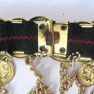 May include: Close-up of a vintage gold-tone belt with black fabric and red stitching. The belt features coin-shaped charms and dangling chains. The clasp is a rectangular gold-tone metal.