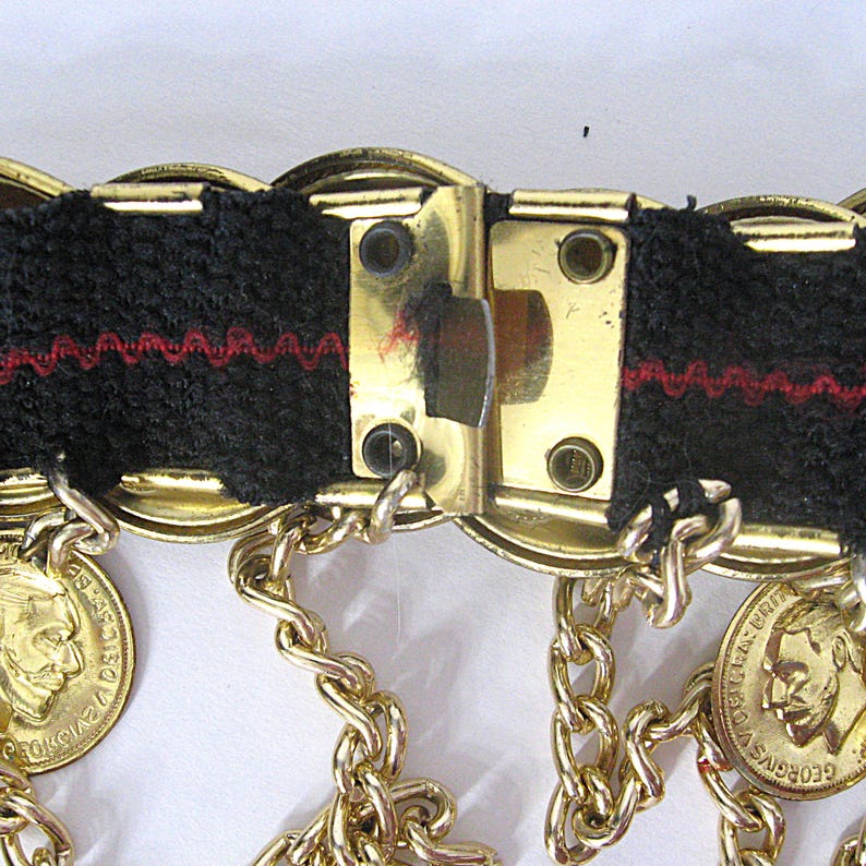 May include: Close-up of a vintage gold-tone belt with black fabric and red stitching. The belt features coin-shaped charms and dangling chains. The clasp is a rectangular gold-tone metal.